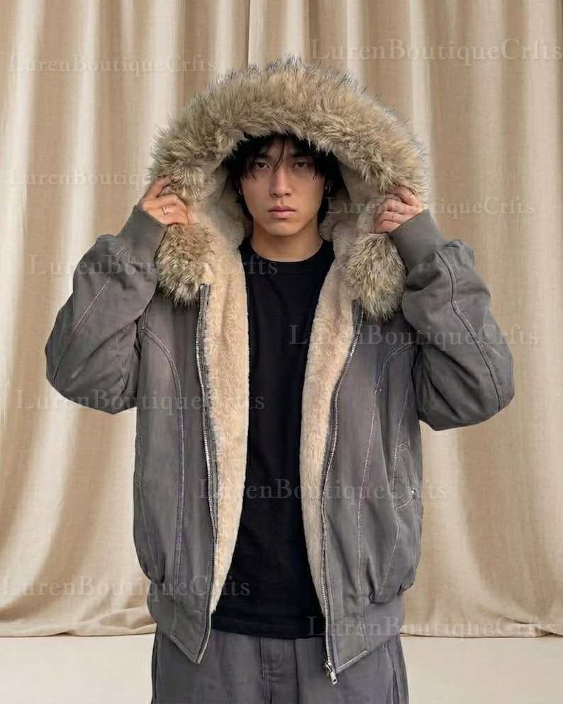 May include: A gray bomber jacket with a faux fur-lined hood. The jacket has a light beige interior lining and a zipper closure. The model is wearing a black shirt underneath. The jacket is a stylish outerwear option.