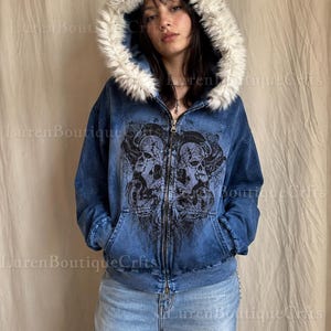 Può includere: Felpa blu con zip e cappuccio foderato in ecopelliccia. La felpa presenta una grafica a teschio nero e due tasche anteriori. La modella indossa jeans azzurri. Da Luren Boutique Crfts.