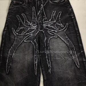 May include: Black denim jeans with a distinctive, distressed design. The jeans showcase a large, stylized spider-like graphic in a lighter grey across the front. The wide-leg style and textured fabric contribute to the edgy aesthetic.