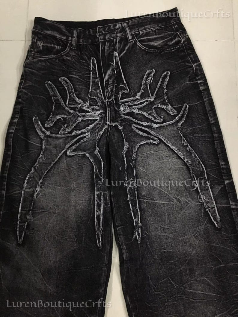May include: Black denim jeans with a distinctive, distressed design. The jeans showcase a large, stylized spider-like graphic in a lighter grey across the front. The wide-leg style and textured fabric contribute to the edgy aesthetic.