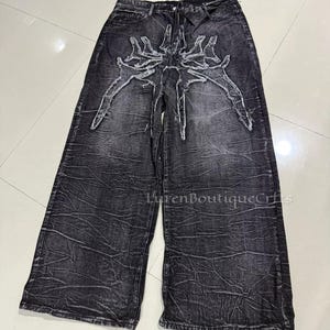 May include: Black denim wide-leg jeans with a distressed texture. The jeans have a white, stylized skeletal design on the front. They have a high waist and a relaxed fit. The jeans are a dark grey colour.