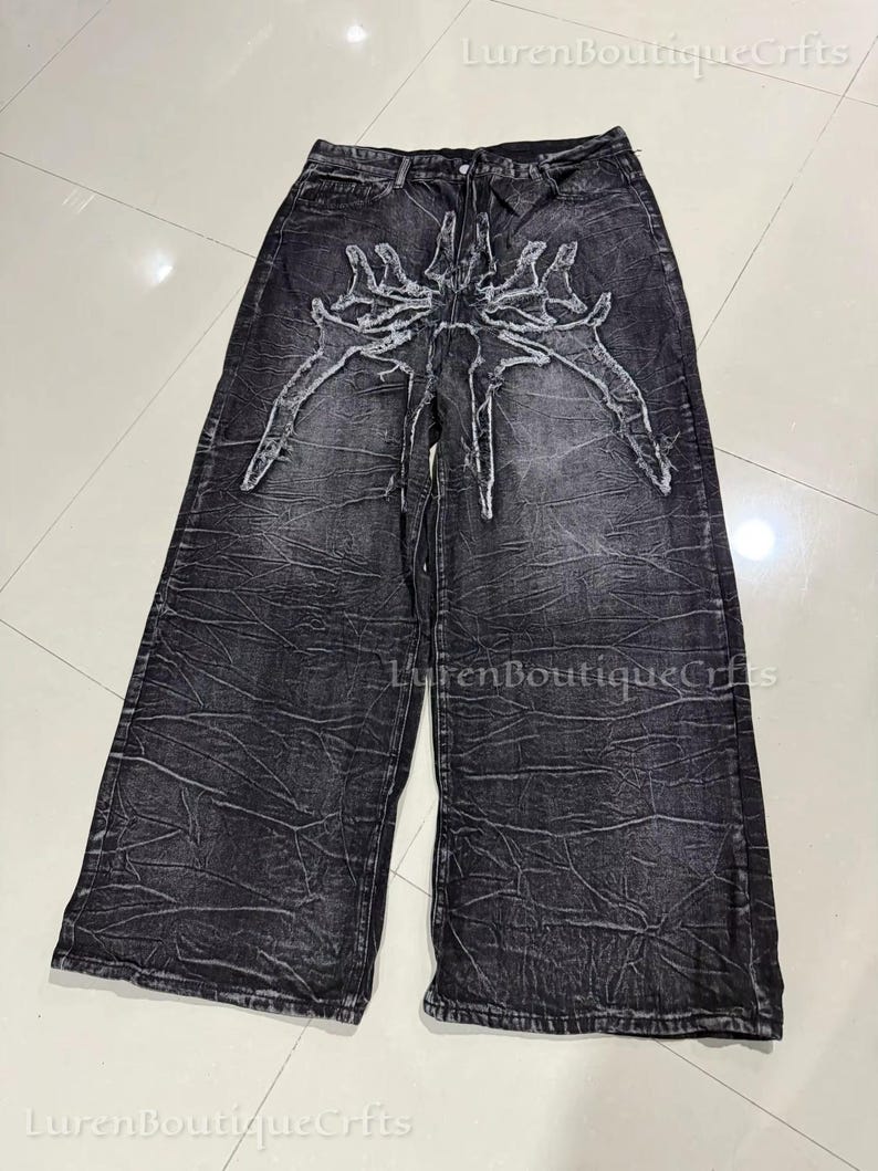 May include: Black denim wide-leg jeans with a distressed texture. The jeans have a white, stylized skeletal design on the front. They have a high waist and a relaxed fit. The jeans are a dark grey colour.