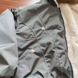 May include: A gray bomber jacket with a fur-lined hood and a white faux fur interior. The jacket has two silver buttons and a zipper closure. The jacket is made of a smooth, water-resistant material.