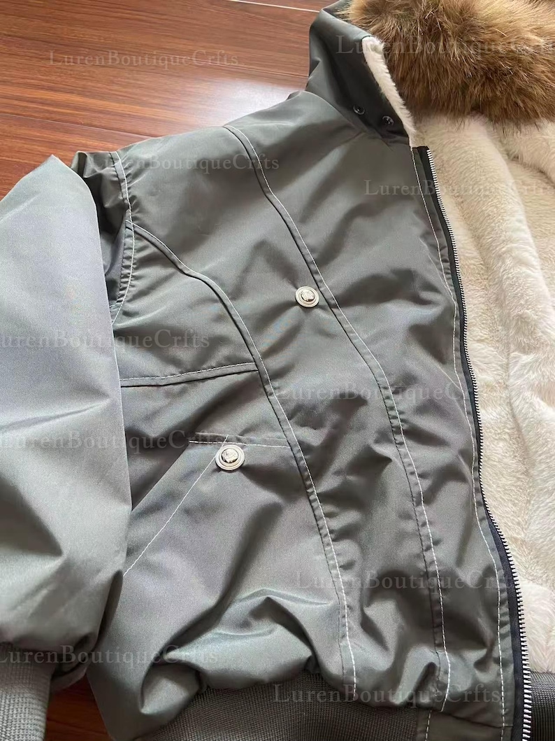 May include: A gray bomber jacket with a fur-lined hood and a white faux fur interior. The jacket has two silver buttons and a zipper closure. The jacket is made of a smooth, water-resistant material.