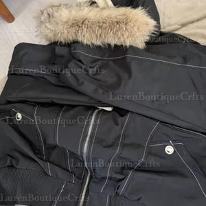May include: Black bomber jacket with a hood trimmed with faux fur. The jacket features white stitching details, a zipper closure, and two front pockets. The hood is lined with a cream-colored material. The jacket has a ribbed hem and cuffs.