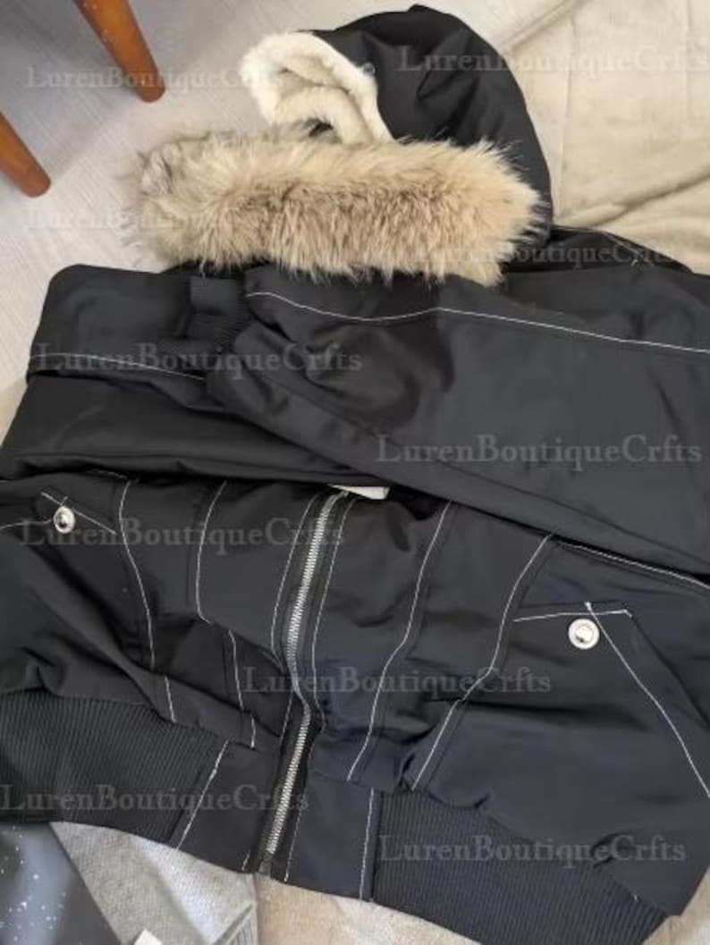 May include: Black bomber jacket with a hood trimmed with faux fur. The jacket features white stitching details, a zipper closure, and two front pockets. The hood is lined with a cream-colored material. The jacket has a ribbed hem and cuffs.