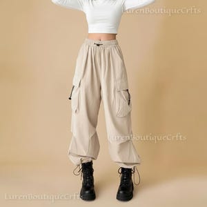 May include: Beige cargo pants with multiple pockets and a drawstring waist. The pants are paired with a white long-sleeve crop top and black combat boots. The pants have a relaxed fit.