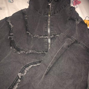 Puede incluir: Chaqueta vaquera gris oscuro con cuello alto y cremallera plateada de largo completo. La chaqueta presenta detalles desgastados a lo largo de las costuras y los bordes, lo que le da un aspecto vintage. La chaqueta est&aacute; hecha de un material vaquero resistente.