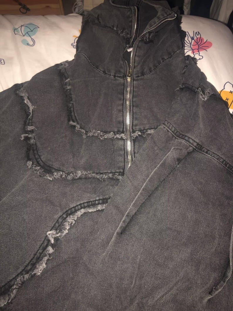 Puede incluir: Chaqueta vaquera gris oscuro con cuello alto y cremallera plateada de largo completo. La chaqueta presenta detalles desgastados a lo largo de las costuras y los bordes, lo que le da un aspecto vintage. La chaqueta est&aacute; hecha de un material vaquero resistente.