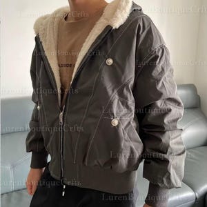 May include: Dark gray hooded jacket with a cream-colored faux fur lining. The jacket features a zip-up front, decorative buttons, and white stitching details. The jacket has a ribbed hem and cuffs.