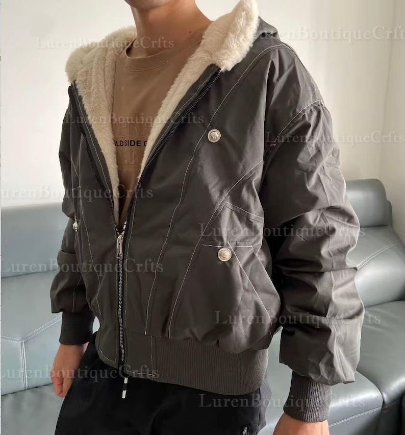 May include: Dark gray hooded jacket with a cream-colored faux fur lining. The jacket features a zip-up front, decorative buttons, and white stitching details. The jacket has a ribbed hem and cuffs.