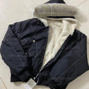 May include: Black hooded jacket with a faux fur-lined hood and a cream-colored, fuzzy interior. The jacket has a zipper closure and a tag attached. White stitching accents the jacket's design.