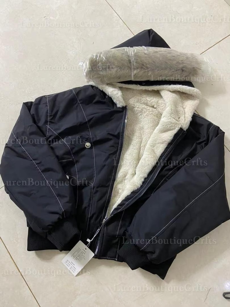 May include: Black hooded jacket with a faux fur-lined hood and a cream-colored, fuzzy interior. The jacket has a zipper closure and a tag attached. White stitching accents the jacket's design.