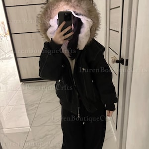 May include: Black winter jacket with a fur-lined hood. The jacket has a zipper closure and is paired with black pants. The image is taken indoors, with a mirror and door visible in the background.