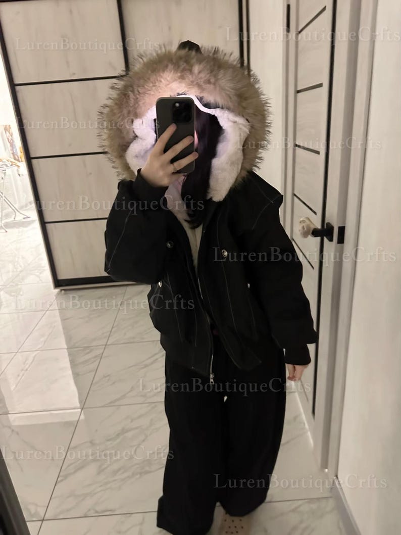 May include: Black winter jacket with a fur-lined hood. The jacket has a zipper closure and is paired with black pants. The image is taken indoors, with a mirror and door visible in the background.