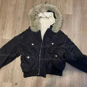 May include: Black bomber jacket with a faux fur-trimmed hood. The jacket features a zip closure, multiple pockets, and white stitching details. The interior lining is white.