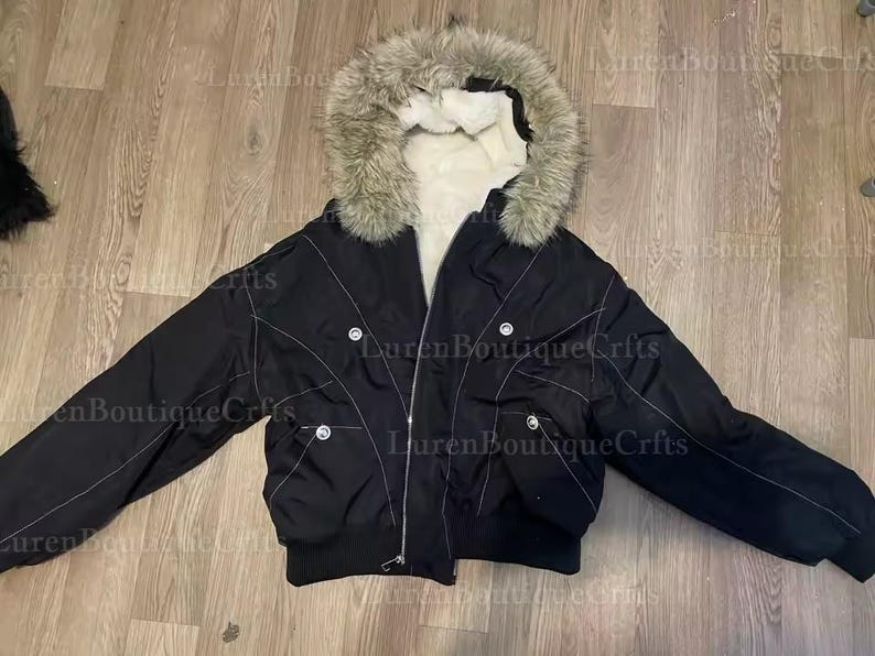 May include: Black bomber jacket with a faux fur-trimmed hood. The jacket features a zip closure, multiple pockets, and white stitching details. The interior lining is white.