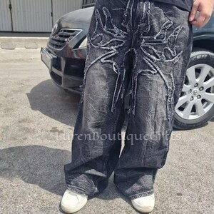 May include: Dark grey denim jeans with a distinctive distressed pattern. The jeans have a large, abstract design in white thread on the front. The text "LurenBoutiqueCfts" is visible in the upper right corner.