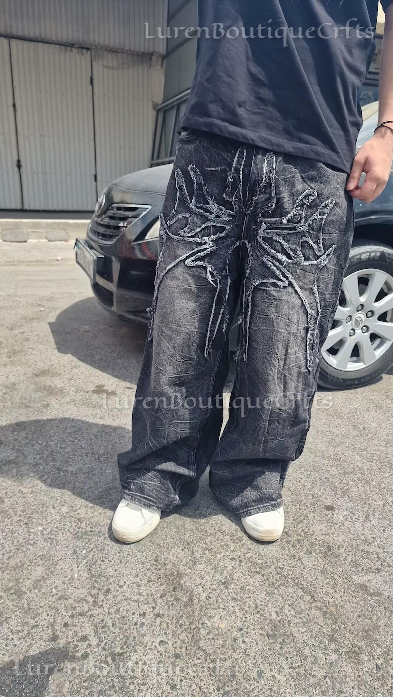 May include: Dark grey denim jeans with a distinctive distressed pattern. The jeans have a large, abstract design in white thread on the front. The text "LurenBoutiqueCfts" is visible in the upper right corner.