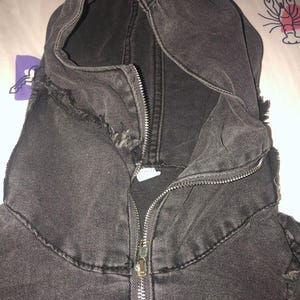 Puede incluir: Primer plano de una chaqueta con capucha de mezclilla negra con cremallera completa. La chaqueta tiene un aspecto desgastado, con bordes deshilachados alrededor de la capucha y las costuras. La tela es de un negro oscuro y descolorido.