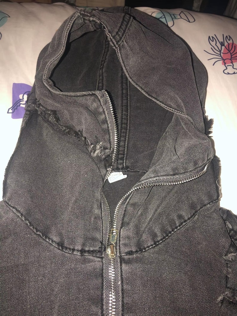 Puede incluir: Primer plano de una chaqueta con capucha de mezclilla negra con cremallera completa. La chaqueta tiene un aspecto desgastado, con bordes deshilachados alrededor de la capucha y las costuras. La tela es de un negro oscuro y descolorido.