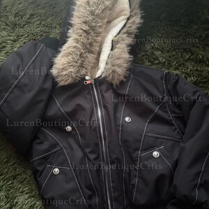 May include: Black bomber jacket with a faux fur-trimmed hood and white stitching details. The jacket features a zipper closure, pockets, and decorative buttons. The hood is lined with a soft, white material.