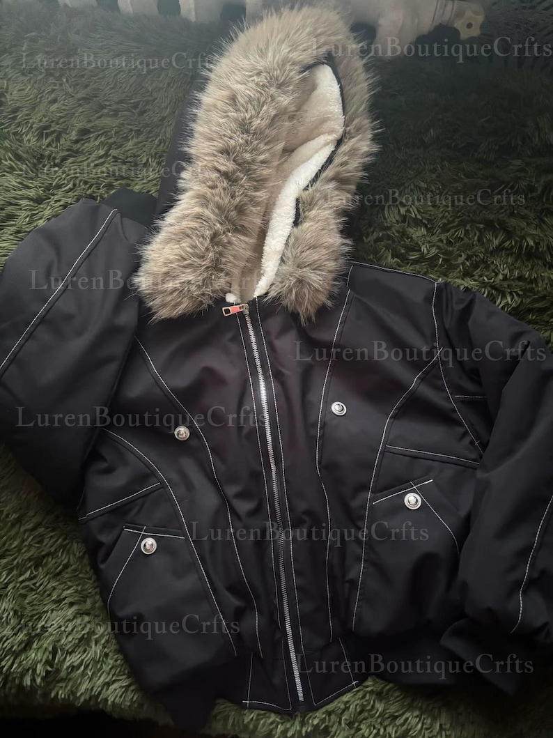 May include: Black bomber jacket with a faux fur-trimmed hood and white stitching details. The jacket features a zipper closure, pockets, and decorative buttons. The hood is lined with a soft, white material.