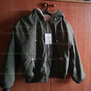 May include: An olive green bomber jacket with a hood and a cream-colored lining. The jacket has a zipper closure, two snap closures, and a tag hanging from the zipper. The jacket is hanging on a black hanger.