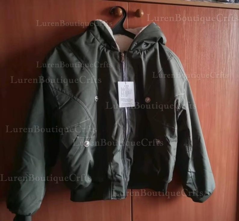 May include: An olive green bomber jacket with a hood and a cream-colored lining. The jacket has a zipper closure, two snap closures, and a tag hanging from the zipper. The jacket is hanging on a black hanger.