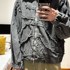 Puede incluir: Chaqueta vaquera gris con cremallera y capucha. La chaqueta presenta detalles desgastados y un dise&ntilde;o &uacute;nico. La persona que sostiene el tel&eacute;fono tiene una funda negra con el texto "MUSIC VM 212UM".