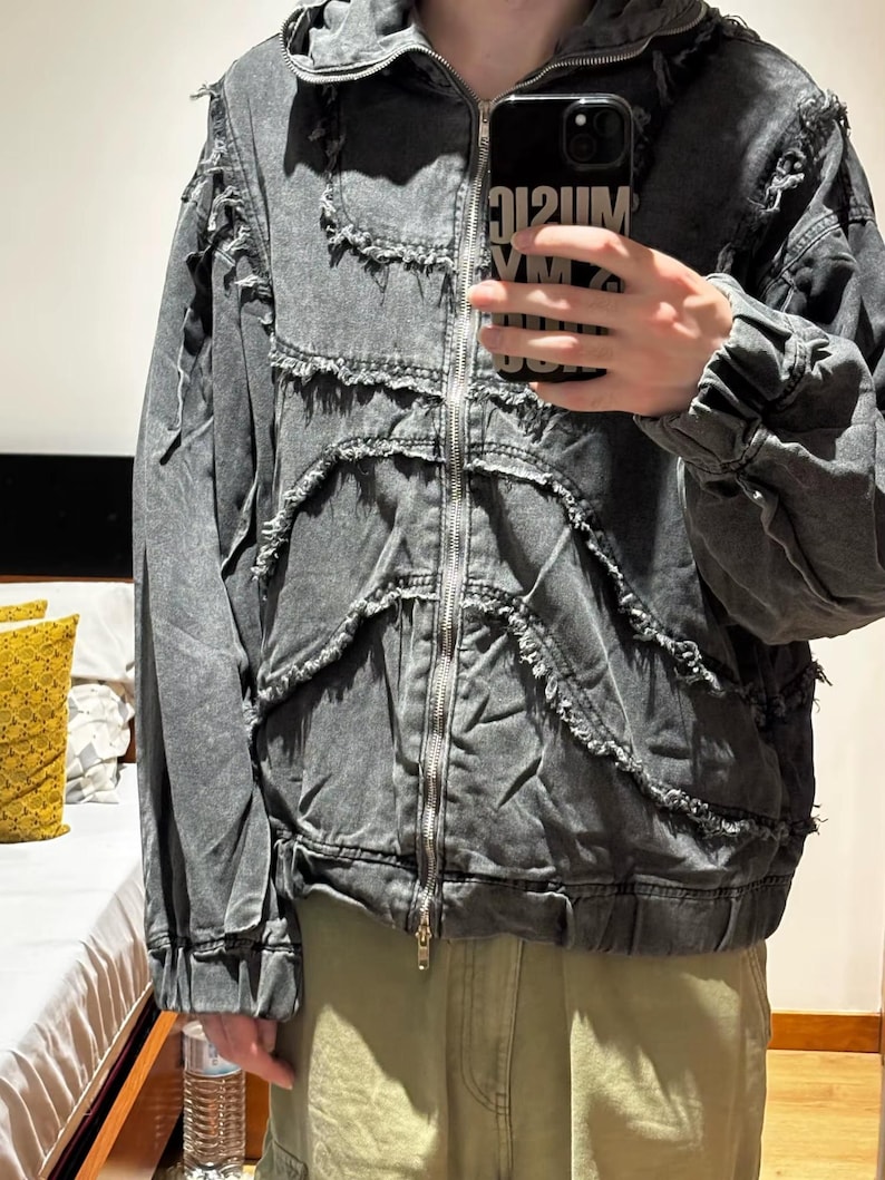 Puede incluir: Chaqueta vaquera gris con cremallera y capucha. La chaqueta presenta detalles desgastados y un dise&ntilde;o &uacute;nico. La persona que sostiene el tel&eacute;fono tiene una funda negra con el texto "MUSIC VM 212UM".