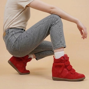 Peut inclure: Baskets montantes rouges avec trois fermetures auto-agrippantes. Les baskets ont un talon compensé et une semelle marron. Les chaussures sont en daim. La personne porte un jean gris et un t-shirt beige.