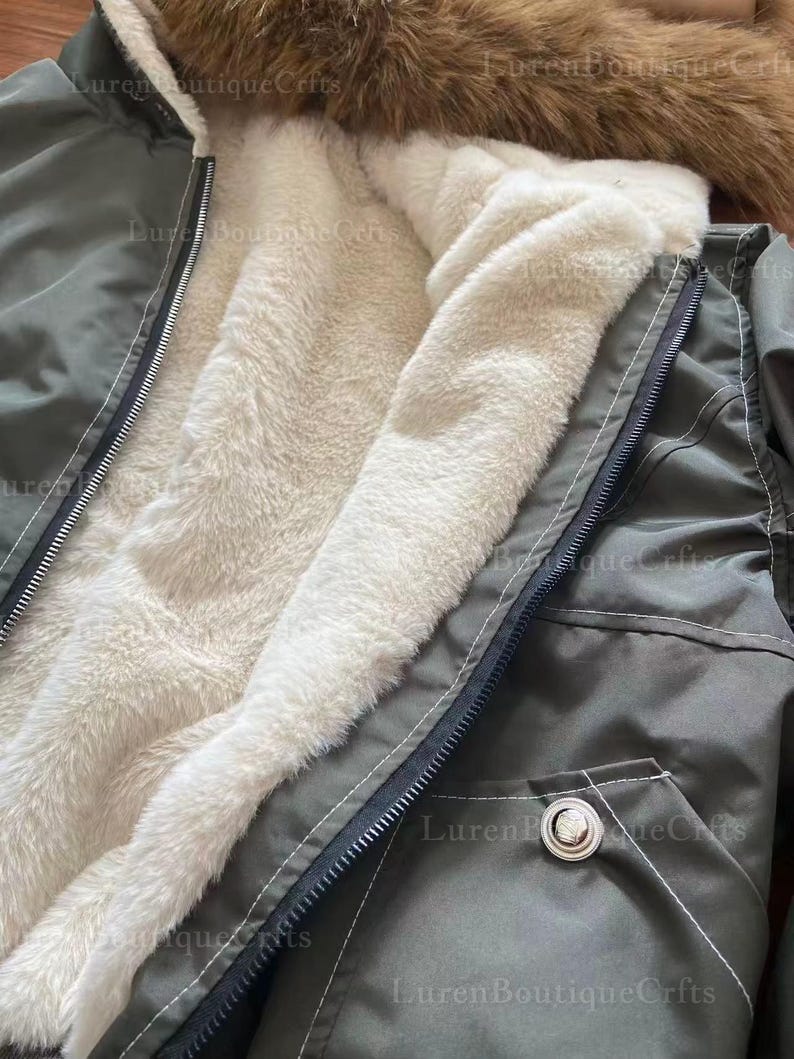 May include: Close-up of an olive green winter coat with a cream-colored, fluffy interior lining. The coat features a black zipper, a buttoned pocket, and a brown fur-lined hood. The image highlights the coat's warm and cozy design.