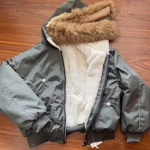 May include: A gray bomber jacket with a hood trimmed with brown faux fur. The jacket has a white, fluffy interior lining and a zipper closure. It features two front pockets and a button detail on the chest.