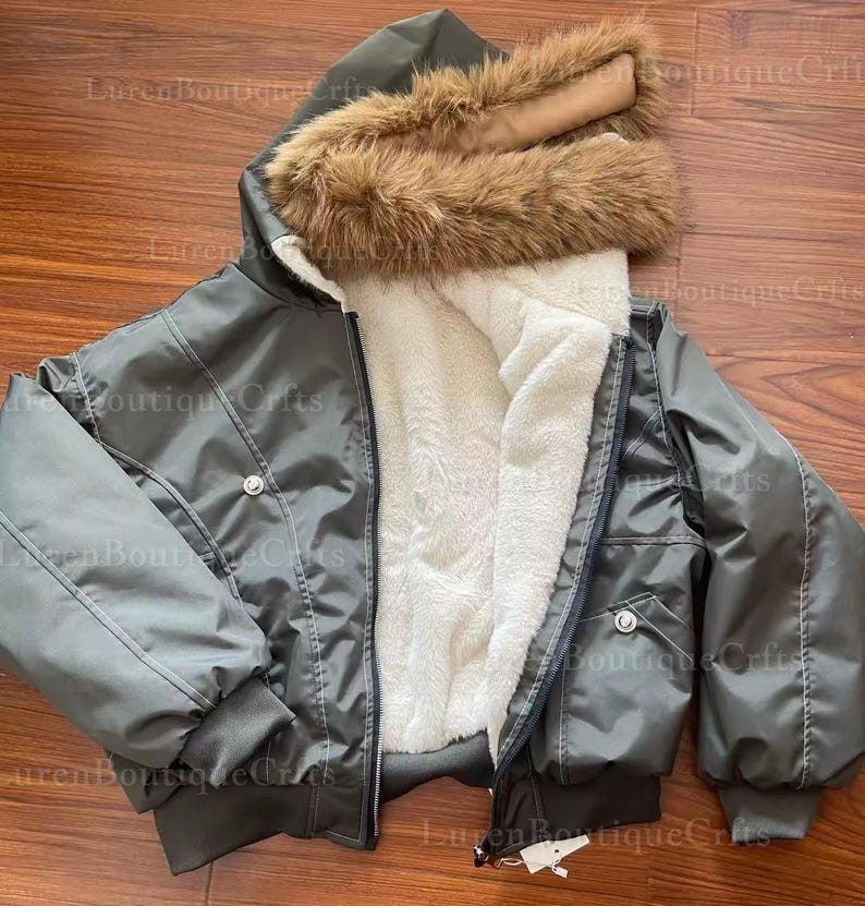 May include: A gray bomber jacket with a hood trimmed with brown faux fur. The jacket has a white, fluffy interior lining and a zipper closure. It features two front pockets and a button detail on the chest.