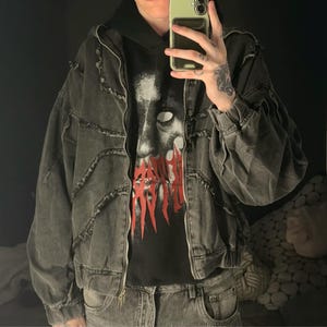 Puede incluir: Chaqueta vaquera gris oscuro con dise&ntilde;o desgastado, cremallera y sudadera con capucha debajo. La sudadera tiene un estampado gr&aacute;fico con texto rojo. La chaqueta tiene un corte holgado.