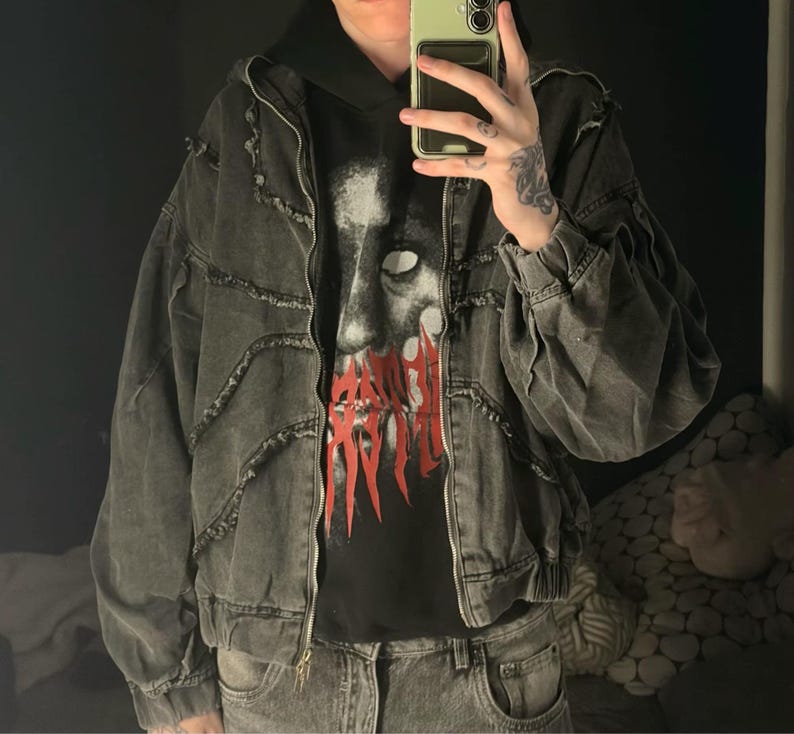 Puede incluir: Chaqueta vaquera gris oscuro con dise&ntilde;o desgastado, cremallera y sudadera con capucha debajo. La sudadera tiene un estampado gr&aacute;fico con texto rojo. La chaqueta tiene un corte holgado.