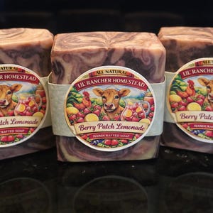 Puede incluir: Tres barras cuadradas de jabón artesanal con un patrón veteado de color púrpura oscuro y marrón. Cada barra tiene una etiqueta redonda con una vaca, fruta y el texto "Berry Patch Lemonade" y "Lil' Rancher Homestead".