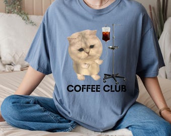 May include: A light blue t-shirt featuring a cartoon cat with an IV drip of coffee. The text "COFFEE CLUB" is printed in black below the cat illustration. The shirt is made of a soft material.