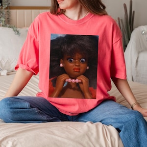 May include: A coral-colored t-shirt featuring a graphic of a doll with a surprised expression. The doll has dark skin, large blue eyes, and is holding a pink pearl necklace. The shirt is paired with blue jeans.
