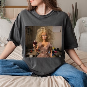 May include: Dark grey t-shirt featuring a framed image of a Barbie doll holding a glass of red wine. The doll has messy blonde hair and wears a pink dress. The shirt is paired with blue jeans.