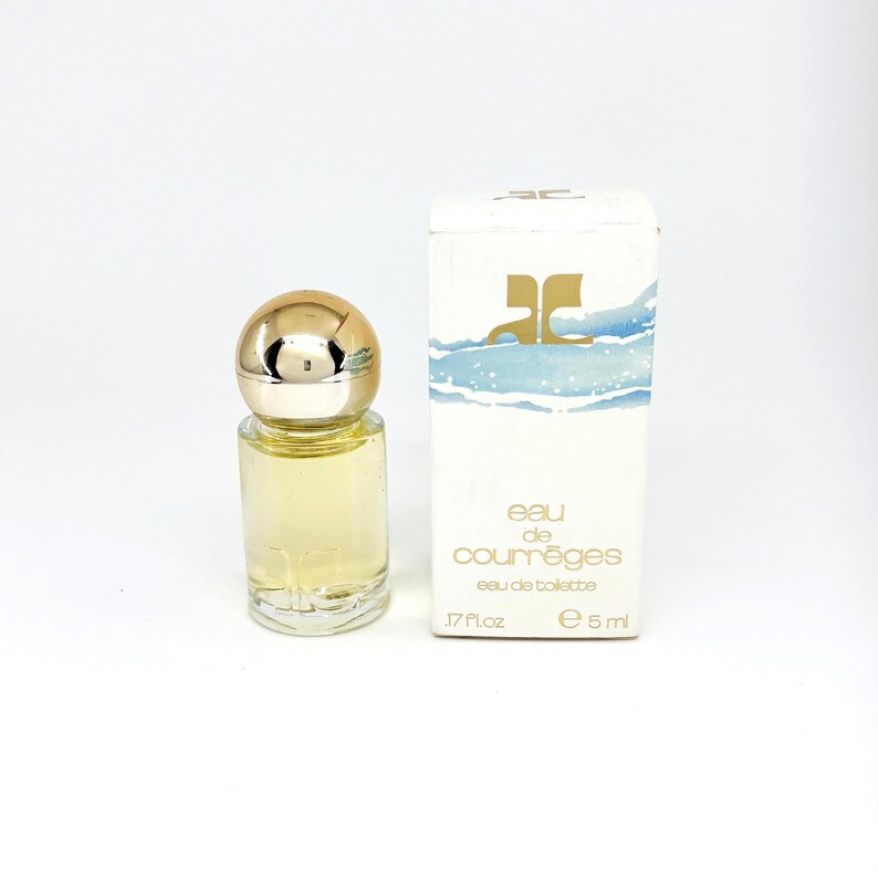 May include: A small perfume bottle with a gold cap and a clear glass body, next to a white box. The box has the words "eau de courr&egrave;ges eau de toilette" printed on it. The bottle contains a light yellow liquid.