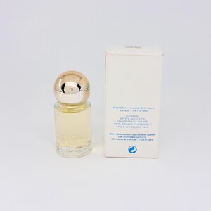 May include: A clear glass perfume bottle with a gold-colored cap and a white box. The bottle contains a light yellow liquid. The box has text that reads "sample - not for sale".