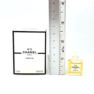 Op de afbeelding: Een witte doos en een kleine Chanel No. 5 parfumfles. De doos heeft zwarte randen en het Chanel-logo. De parfumfles is ongeveer 4 cm hoog. Een liniaal staat naast de doos en de fles.