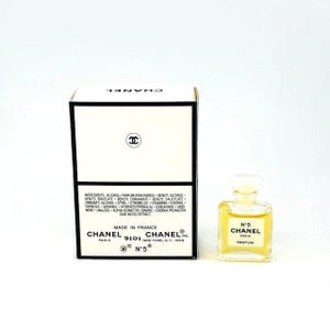 Op de afbeelding: Een kleine fles Chanel No. 5 parfum en de doos. De doos is wit met zwarte randen en het Chanel-logo. De parfumfles is helder, bevat een goudkleurige vloeistof en heeft een wit etiket met "N&deg;5 CHANEL PARFUM".