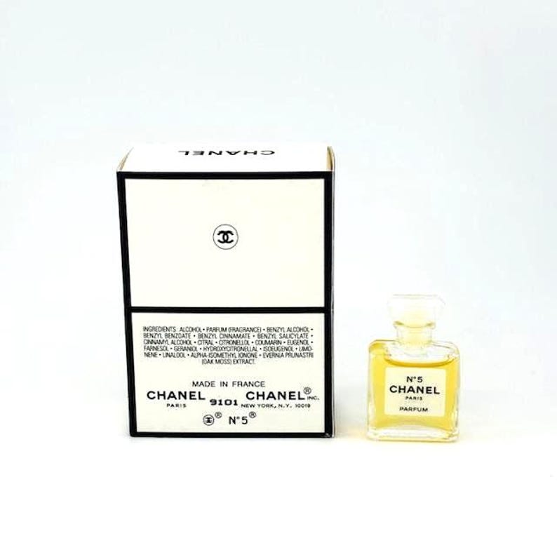 Op de afbeelding: Een kleine fles Chanel No. 5 parfum en de doos. De doos is wit met zwarte randen en het Chanel-logo. De parfumfles is helder, bevat een goudkleurige vloeistof en heeft een wit etiket met "N&deg;5 CHANEL PARFUM".