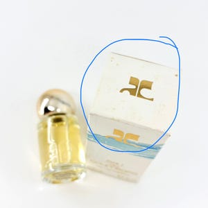 May include: A miniature perfume bottle with a silver cap and a clear glass body filled with a yellow liquid. Beside it is a white box with a gold logo and blue accents. The box has the logo "JC" on it.