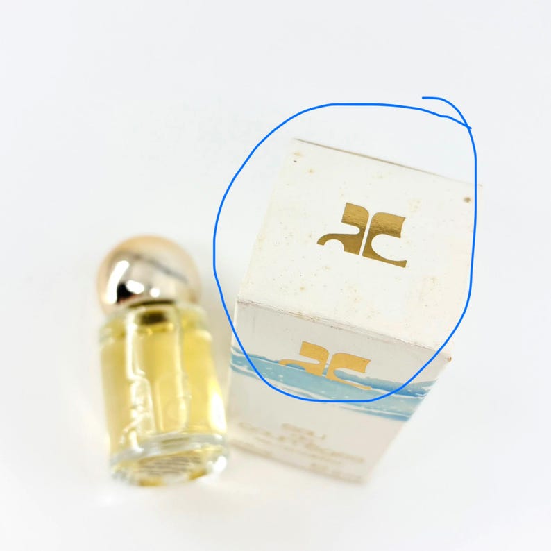 May include: A miniature perfume bottle with a silver cap and a clear glass body filled with a yellow liquid. Beside it is a white box with a gold logo and blue accents. The box has the logo "JC" on it.