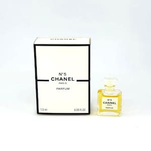 Op de afbeelding: Een kleine fles Chanel No. 5 Parfum naast een witte doos met zwarte tekst. Zowel de doos als de fles tonen "N&deg;5 CHANEL PARIS PARFUM". De fles bevat een gele vloeistof. De doos is gemarkeerd met 1,5 ml en 0,05 fl oz.