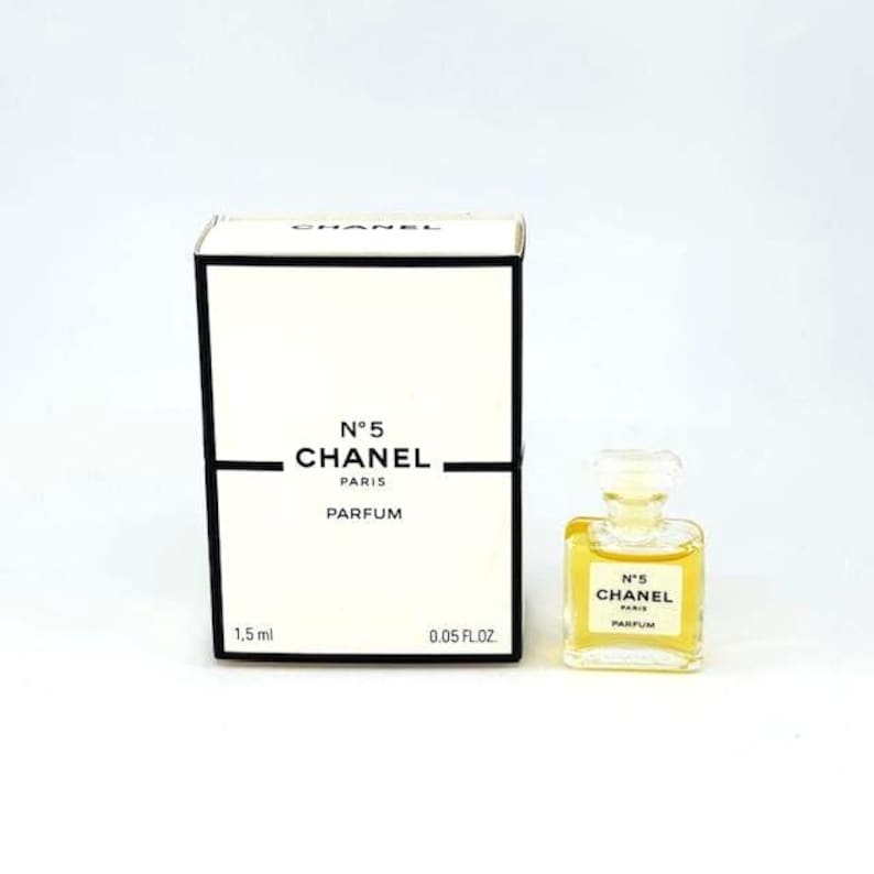 Op de afbeelding: Een kleine fles Chanel No. 5 Parfum naast een witte doos met zwarte tekst. Zowel de doos als de fles tonen "N&deg;5 CHANEL PARIS PARFUM". De fles bevat een gele vloeistof. De doos is gemarkeerd met 1,5 ml en 0,05 fl oz.